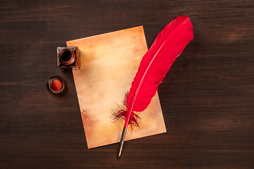 A vintage background with a vibrant red quill on a piece of old paper, with an ink well, on a dark wooden background with copy space