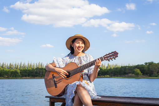 Happy young Asian woman playing classical guitar at lakeside and blue sky in background