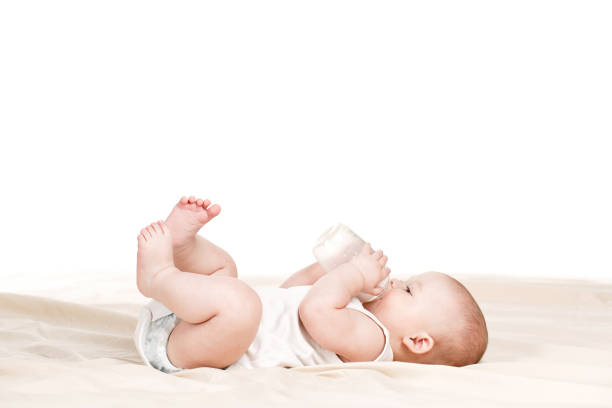 Baby drinking milk in white crib.Infant boy with formula bottle in bed. Kids nutrition.Healthy food and drink for children.Child in white nursery.Baby room interior.Kid with toy and milk bottle.Pretty baby boy drinks water from bottle lying on bed.