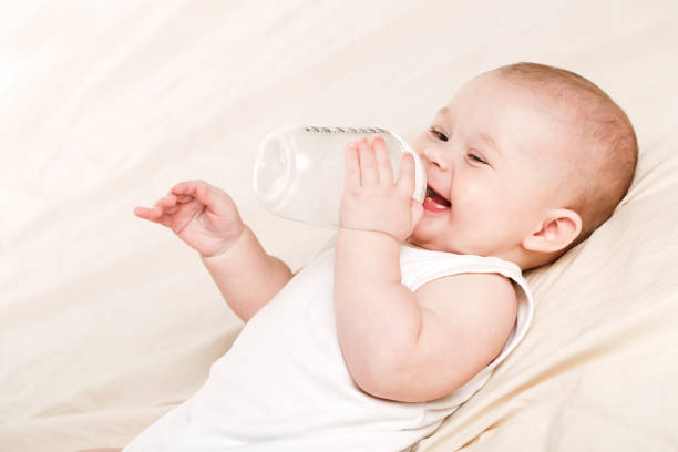 Baby drinking milk in white crib.Infant boy with formula bottle in bed. Kids nutrition.Healthy food and drink for children.Child in white nursery.Baby room interior.Kid with toy and milk bottle.Pretty baby boy drinks water from bottle lying on bed.