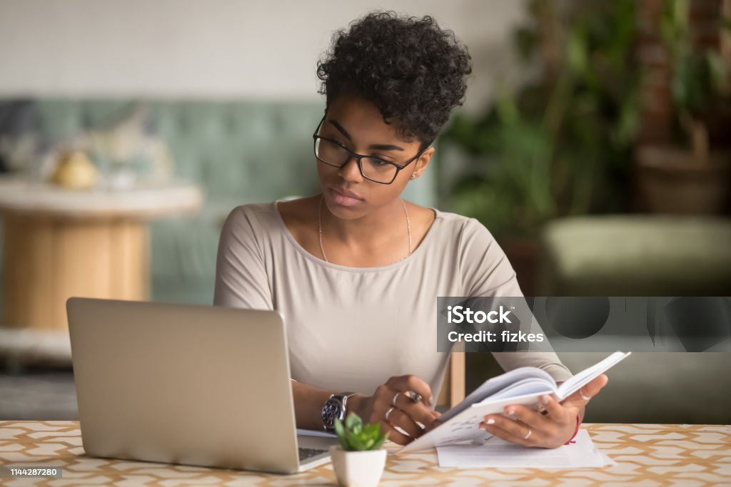 Focused african student looking at laptop holding book doing research Focused young african american businesswoman or student looking at laptop holding book learning, serious black woman working or studying with computer doing research or preparing for exam online Learning Stock Photo