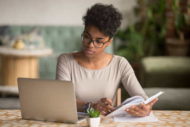 Focused african student looking at laptop holding book doing research Focused young african american businesswoman or student looking at laptop holding book learning, serious black woman working or studying with computer doing research or preparing for exam online homework stock pictures, royalty-free photos & images