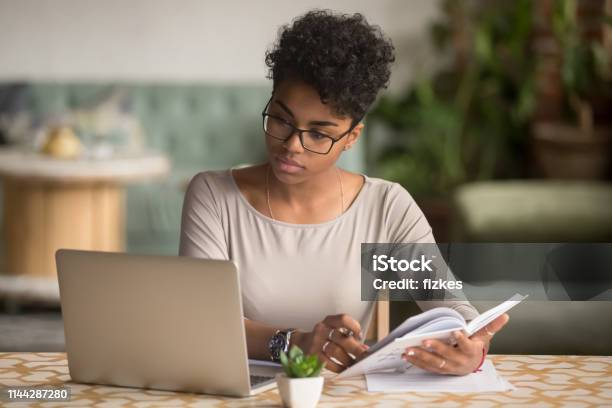 Focused African Student Looking At Laptop Holding Book Doing Research Stock Photo - Download Image Now