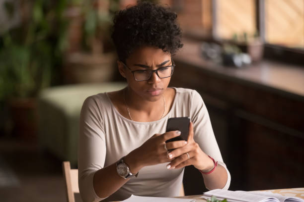 Upset confused african woman holding cellphone having problem with phone Upset confused african woman holding cellphone having problem with mobile phone, frustrated angry mixed race girl reading bad news in message looking at smartphone annoyed by spam or missed call white collar crime stock pictures, royalty-free photos & images