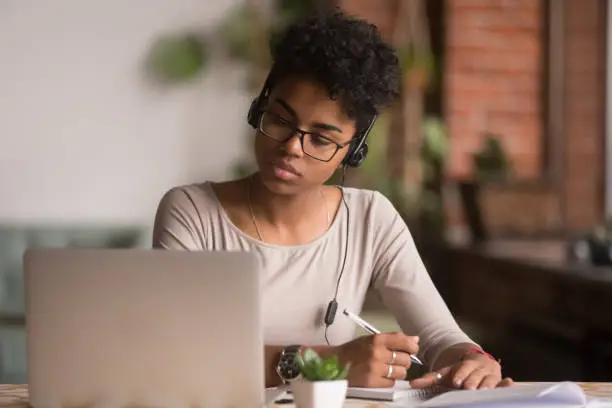 Photo of Focused mixed race woman wearing headphones watching webinar write notes