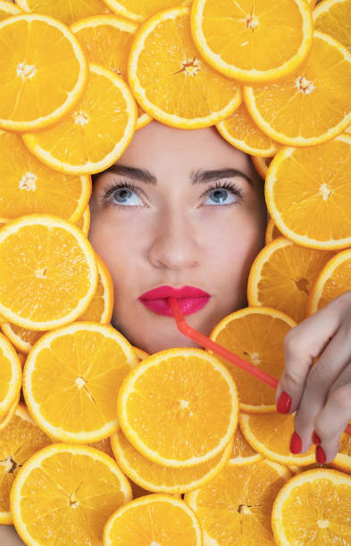 Charming woman with bright lipstick and orange slices on her face drinking juice through a red straw stock photo