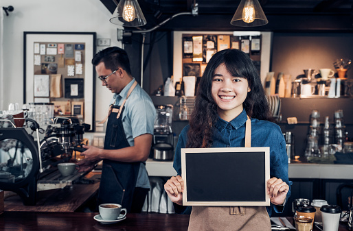 Asian female barista wear jean apron holding blank blackboard coffee menu at counter bar with colleague with smile face ,cafe service concept,business owner start upLeave space for adding your text.
