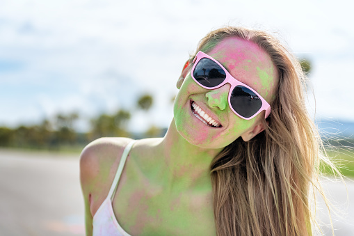 Woman loving the color festival, laughing and looking at camera outdoors. Young candid girl with face paint wearing pink sunglasses and enjoying the event.