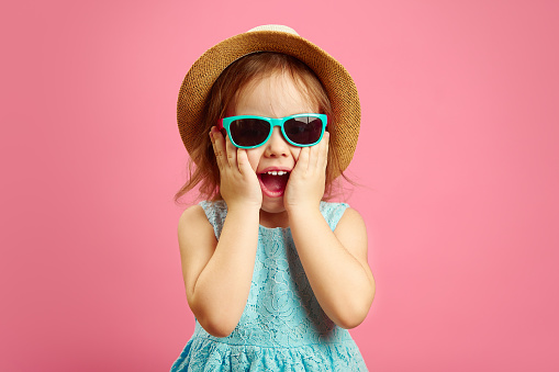 Portrait of surprised girl with open moutn, wears in panama hat and sunglasses, expresses surprise and delight, stands over pink isolated background. Happy child is looking forward to vacation.