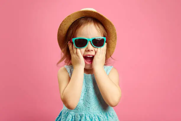Photo of Portrait of surprised girl with open moutn, wears in panama hat and sunglasses, expresses surprise and delight, stands over pink isolated background.