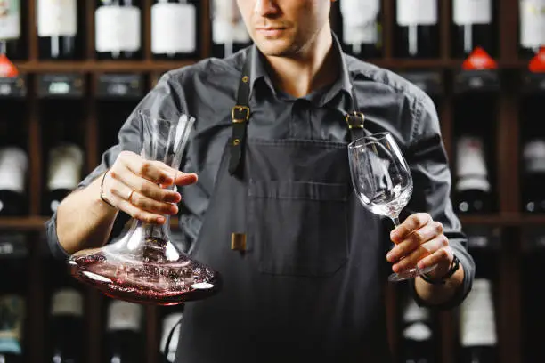 Barman in apron holds big decanter with red wine on bottom and glass, stands in cellar among sealed bottles of alcohol drinks on large wooden shelves.