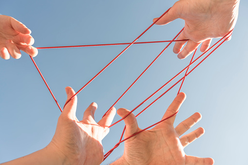 Three adult and one child's hands playing cats cradle against clear blue sky.