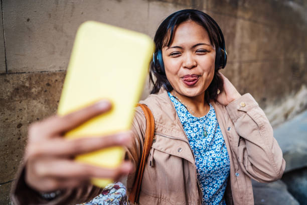 mujer adulta joven tomando un selfie y usando auriculares en la ciudad - filipino ethnicity audio fotografías e imágenes de stock