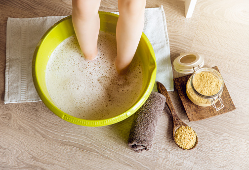 Child taking a healing warming foot bath with mustard powder, adding mustard powder to foot bath with wooden spoon. Against cold illness, aches and improves blood circulation. Alternative medicine.