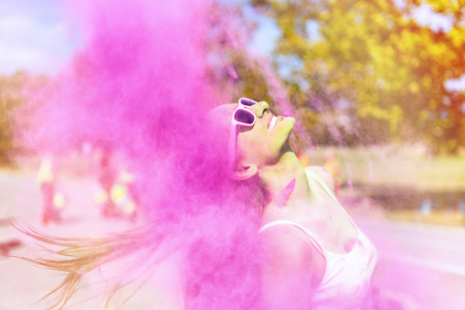 Woman loving the color festival, laughing and splashing hair in pink colored powder. Young candid girl with face paint wearing pink sunglasses and enjoying the event.