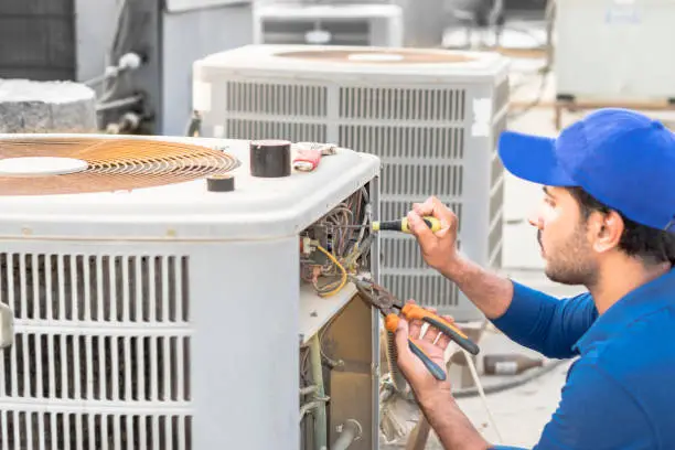 Photo of a professional electrician man is fixing the heavy unit of an air conditioner at the roof top of a building and wearing blue uniform and head cap