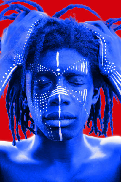 istock A striking dark skinned male shows off traditional white face painted patterns which contrast his skin Portrait of a dark skinned man with dreadlocks and tribal face paint holds hair with his hands. Blue hue on red background. 1144330452