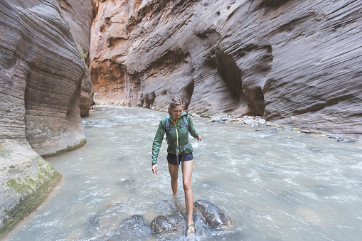 Solo female hiker wades through a majestic red rock canyon in Utah.  The walls are rising up all around her as she steps over large rocks in the water.