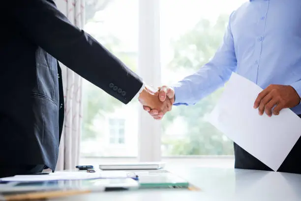 Young applicant with resume and his new employer in formalwear shaking hands over desk on background of office window