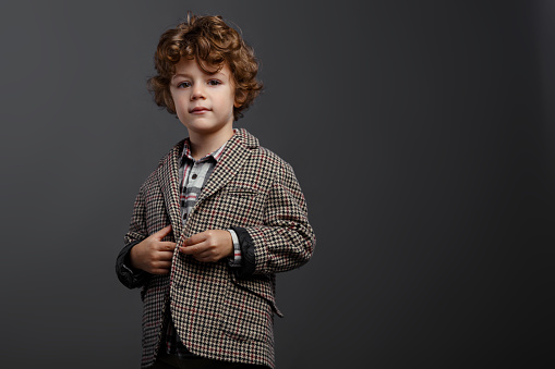 Contented boy portrait of a cute schoolboy with reddish curly hair dressed in a t-shirt and suit, looking at camera, shoting at the studio, isolated on a grey background, with copy space.