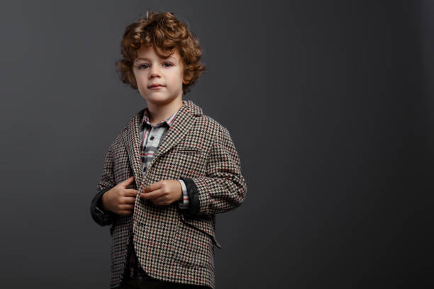 Contented boy cute boy with red-brown curly hair dressed in a elegant suit at the studio, isolated on a grey background, with copy space. stock photo