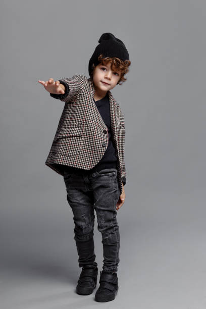 Adorable little boy curly haired, wearing fashionable, raised his right hand forward, isolated on a grey background, looking at camera. stock photo