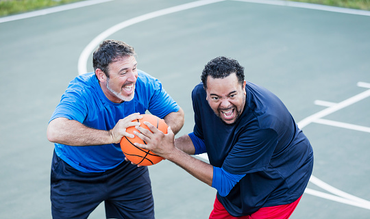 Two mature multi-ethnic men in their 40s having fun playing basketball on an outdoor court. They are having a friendly fight over the ball. The one on the left is Hispanic and his friend is African-American.