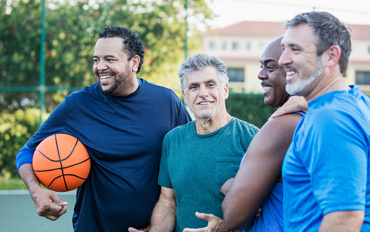 A multi-ethnic group of middle-aged and senior men on an outdoor basketball court. The Caucasian man wearing the green shirt, looking at the camera, is a senior, in his 60s. The bald African-American man is in his 50s, and their friends are mature men in their 40s.