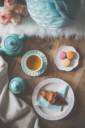 Croissant on the plate, macaroons and tea for breakfast, Flat lay setting of teapot, tea cup, croissant on a plate, macaroons, bouquet of roses and party decor.