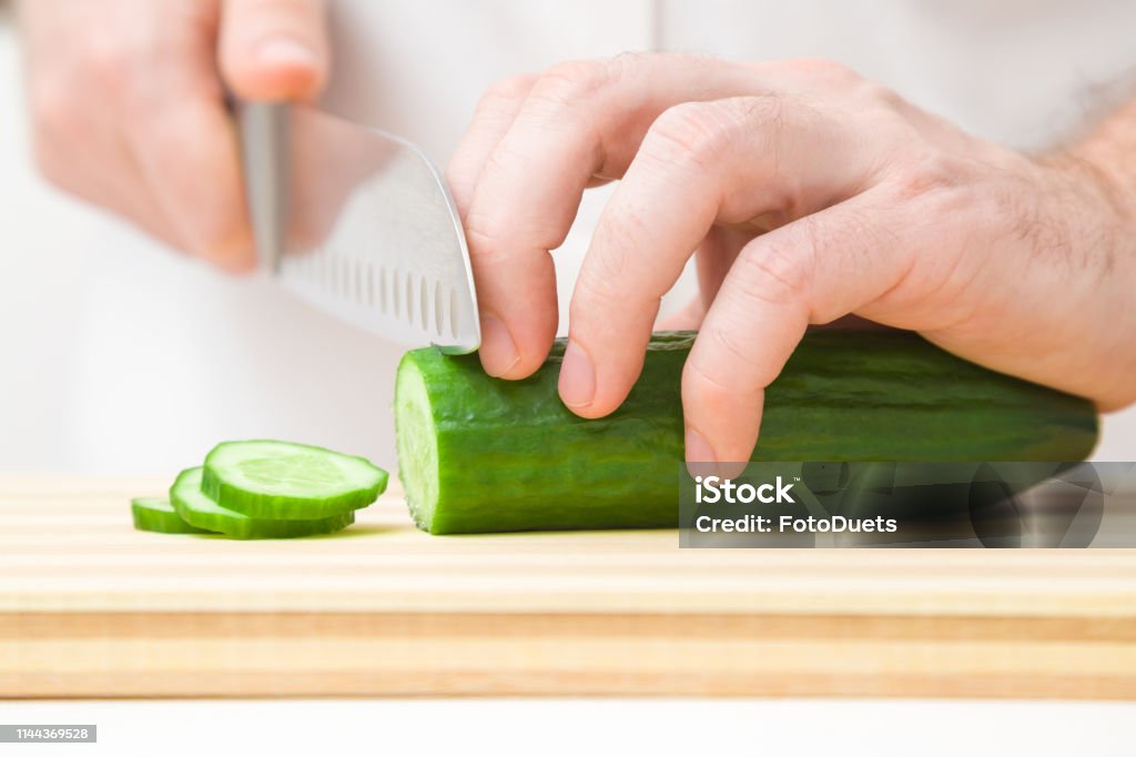 Die Hände des Mannes, die grüne Gurke mit großem Messer auf Holzbrett schneiden. Zutaten für Salat zubereiten. Closeup. Frontansicht. - Lizenzfrei Abnehmen Stock-Foto