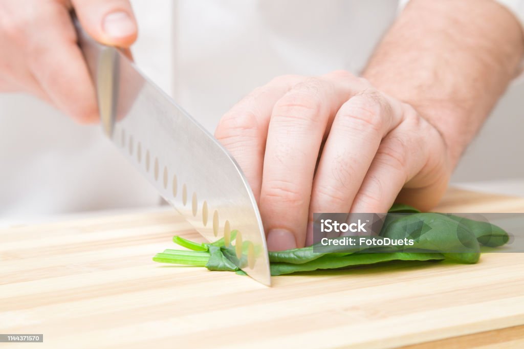 Die Hände des Mannes schneiden grüne Spinatblätter mit einem großen Messer auf Holzbrett. Closeup. Frontansicht. - Lizenzfrei Abnehmen Stock-Foto