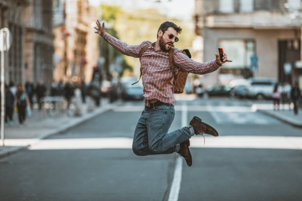 Handsome Man in Plaid Shirt and Sunglasses is Walking in the City Street and Having a Fun. Young Male Tourist with Backpack is Jumping on a Line of the City Road with his Arms Raised nad Holding a Mobile Phone.