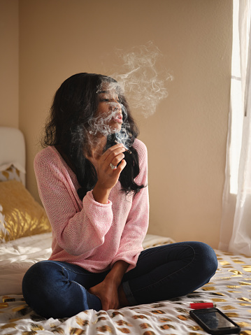 african american woman smoking marijuana joint while sitting on bed at home during the day