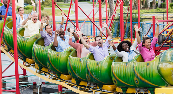 A multi-ethnic group of nine adults at an amusement park participating in corporate team building activities.  The coworkers range in age from 20s to 60s. They are sitting on a small rollercoaster, laughing and shouting with their arms raised over their heads.