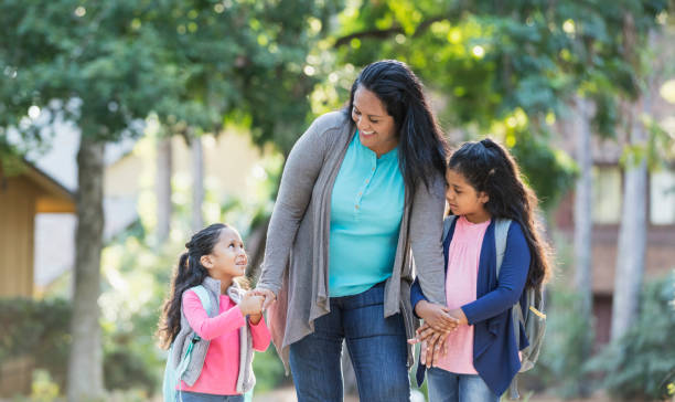 Hispanic mother taking her two girls to school A mature Hispanic woman in her 40s walking with her two young daughters, 4 and 6 years old, holding their hands. The children are carrying backpacks on their way to school. The mother is smiling, looking down at the younger girl who looks a little worried. nervous holding hands stock pictures, royalty-free photos & images