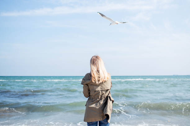 ビーチには女の子が立っており、近くでカモメが飛んでいます。海のビーチで晴れた日。瞬間をお楽しみください。海に休息とリラクゼーション。自然の中で週末。ライフ スタイル - sunny day sunlight seagull ストックフォトと画像