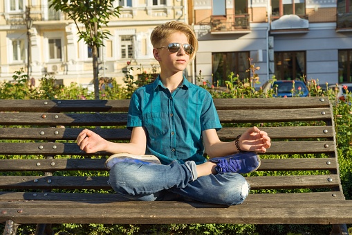 Yoga in city, teenage boy sits in lotus pose on bench in city park. Relax, rest, meditation