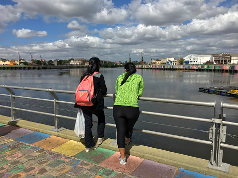 Buenos Aires, Argentina - March 24, 2019: Two women lying on railing while talking to each other facing the Riachuelo river in La Boca neighborhood. This area next to the port is full of colorful houses painted with vibrant colors, an heritage of the first italian immigrants that came from Genoa to live in the city