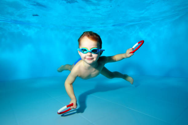 lycklig liten pojke simma under vattnet i poolen, leende och poserar för kameran med leksaker i händerna på en blå bakgrund. porträtt. undervattens fotografering. horisontell orientering - baby swim under water bildbanksfoton och bilder