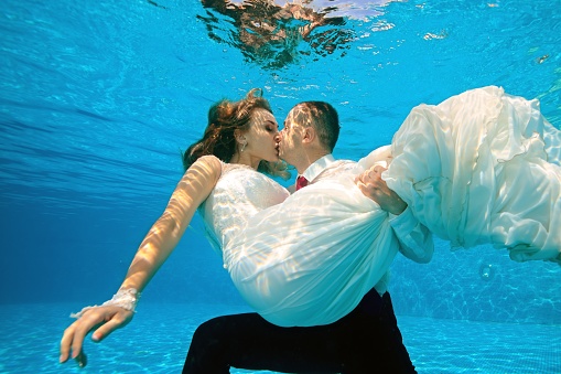 Happy bride and groom kissing underwater in the pool on a Sunny day. The groom hugs and holds the bride in his arms at the bottom. Portrait. Concept. Wedding underwater. Horizontal view.
