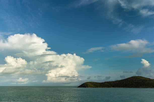 Thursday Island, Australia - February 20, 2019: Wide shot of dark green rainforets covered Goods Island, part of Torres Strait Islands Archipelago under white cloudscape in blue sky. Green-blue sea water in front.