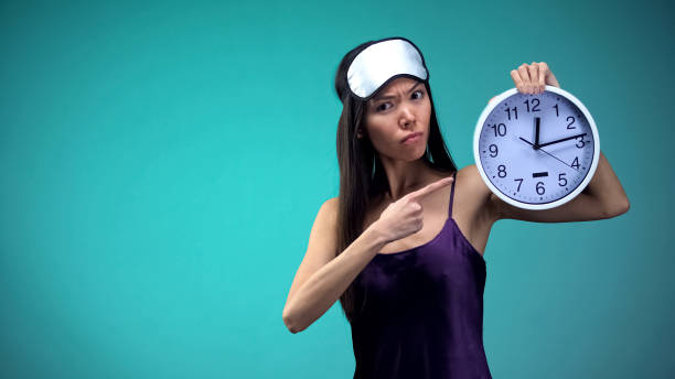 istock Sleepless woman in pajamas and with blindfold on face pointing at clock insomnia 1144432917
