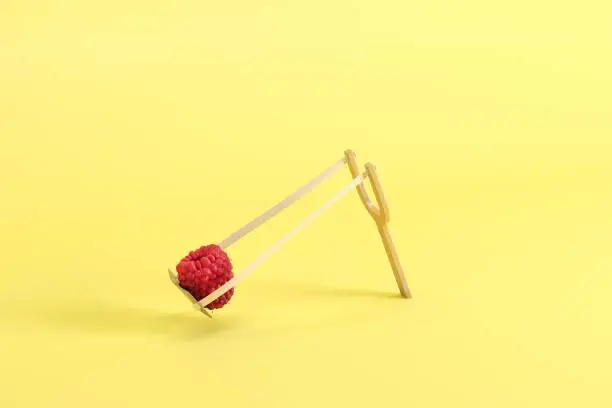 Photo of Raspberry in a slingshot on yellow background. Minimal fruit idea concept.