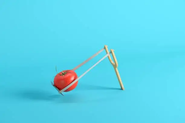 Photo of Fresh red tomato with water drops in a slingshot on blue background. Minimal fruit idea concept.