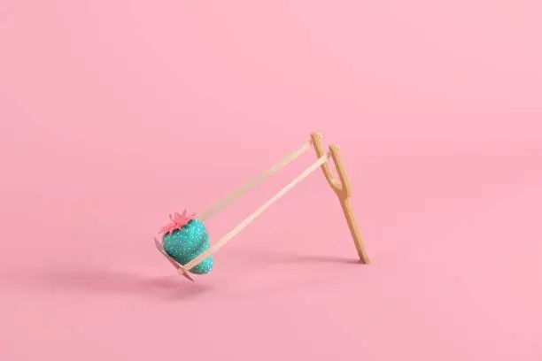 Photo of Strawberry painted in blue with pink leaves and stem in a slingshot on pink background. Minimal fruit idea concept.