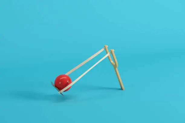 Photo of Red ball in a slingshot on blue background. Minimal fruit idea concept.