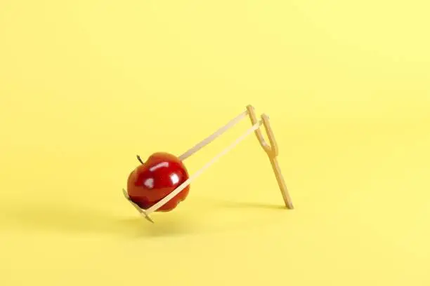 Photo of Red apple in a slingshot on yellow background. Minimal fruit idea concept.