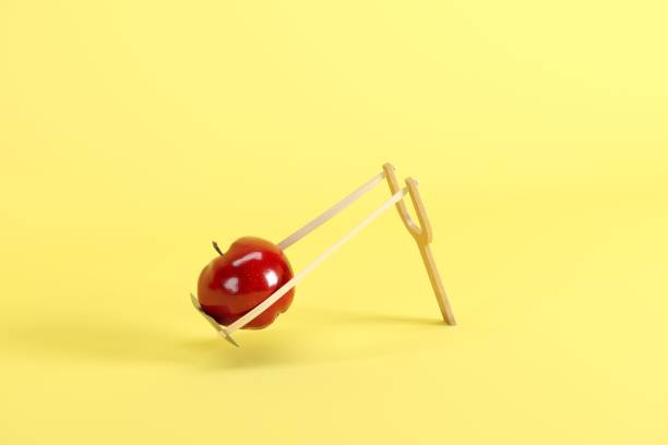 red apple in a slingshot on yellow background. minimal fruit idea concept. - apple fruit surreal bizarre imagens e fotografias de stock