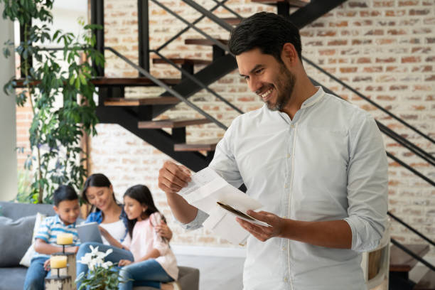 istock Handsome man reading a letter he just received in the mail while spouse and kids are sitting at background playing on tablet 1144447575