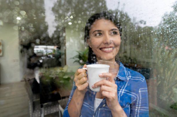 Beautiful woman at home looking through the window on a rainy day enjoyng a cup of coffee Beautiful woman at home looking through the window on a rainy day enjoyng a cup of coffee smiling looking through an object stock pictures, royalty-free photos & images